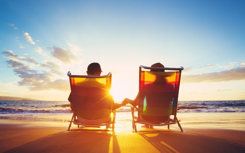 couple sitting on beach in beach chairs holding hands watching the sunset