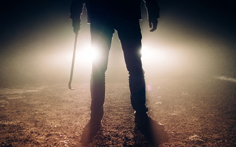 the bottom half of a person holding a crowbar in dark pants backlist by headlights